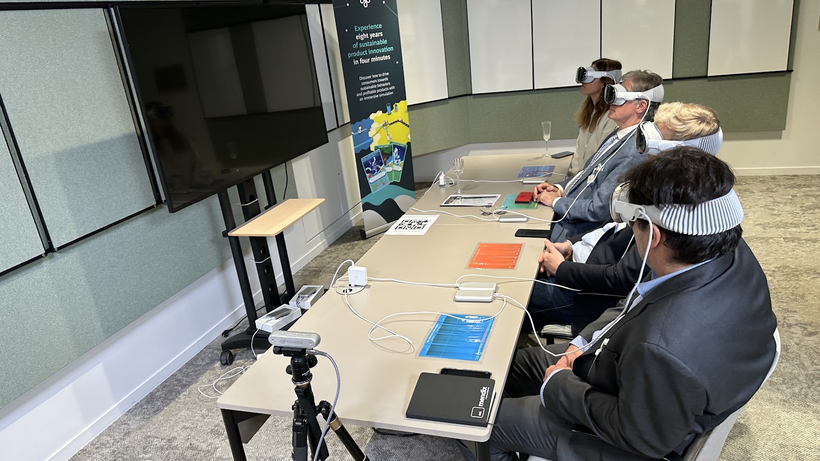 Quatre participants alignés le long d’une table de réunion, chacun équipé d’un casque Apple Vision Pro ; un kakémono annonce « eight years of sustainable product innovation in four minutes » et une caméra sur trépied filme la session.
