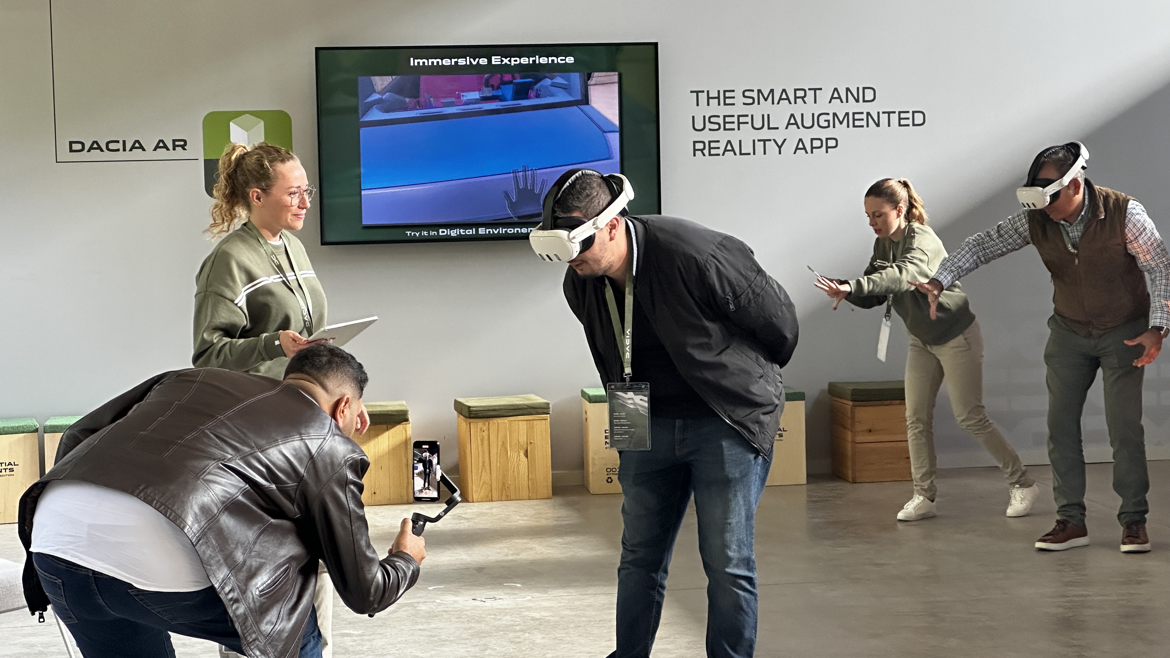 L’espace de démonstration Dacia AR : un participant en casque Meta Quest 3 se penche vers le véhicule virtuel pendant qu’un vidéaste le filme au smartphone ; à droite, deux autres visiteurs tendent les mains vers la voiture.