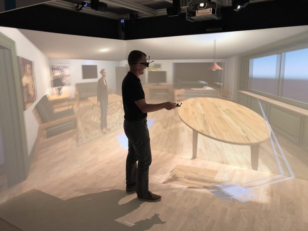Salle immersive à quatre faces de l'ENSIIE : un utilisateur portant des lunettes 3D suivies et tenant une manette se tient dans un séjour virtuel projeté sur les murs et le sol.