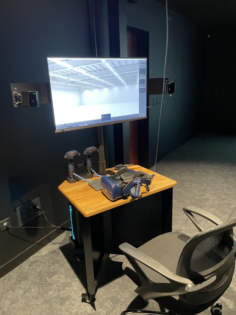 Borne de réalité virtuelle mobile : un casque HTC Vive Pro Eye et deux manettes posés sur un plan de travail en bois, sous un écran de retour affichant un entrepôt en 3D.